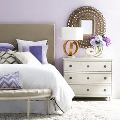 a white bed sitting next to a dresser with purple and white pillows on top of it