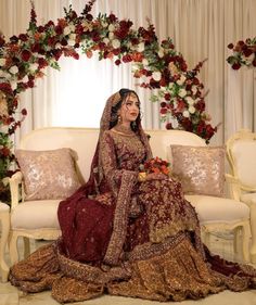 a woman sitting on top of a couch wearing a red and gold wedding dress in front of a floral arch