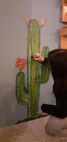 a woman kneeling down next to a painting of a cactus and flowers on the wall