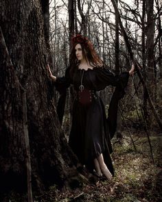 a woman in a black dress standing next to a tree with her hands on her hips