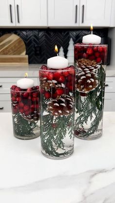 three candles with pine cones and berries in them are sitting on a counter top next to some white cabinets