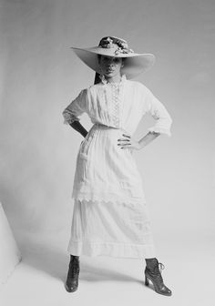 a woman in a white dress and large hat poses for the camera with her hands on her hips