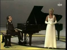 a woman in white dress standing next to a man in black suit at a piano