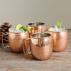 four copper mugs sitting next to each other on top of a wooden table with pine cones