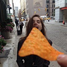 a woman holding up a piece of pizza to her mouth while standing on the sidewalk