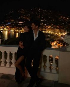 a man and woman standing next to each other on a balcony with the city lights in the background