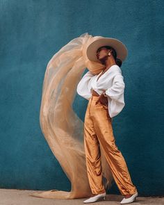 a woman standing in front of a blue wall wearing a white shirt and gold pants