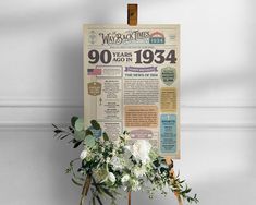 an old newspaper with flowers and greenery on it hanging from a wooden easel