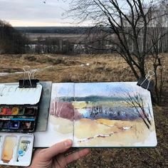 a person holding an open book with watercolors on it and trees in the background