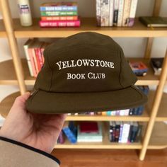 a person holding up a yellow stone book club hat in front of a bookshelf