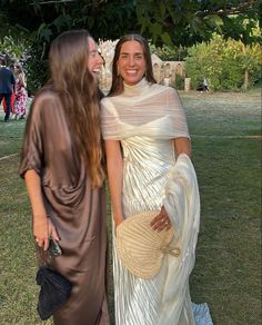 two women standing next to each other in front of trees and grass, one wearing a white dress