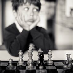 a young boy is playing chess with his hands on his face and looking at the camera