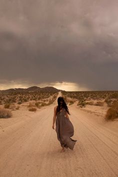 a woman walking down a dirt road in the middle of an open desert area with storm clouds overhead