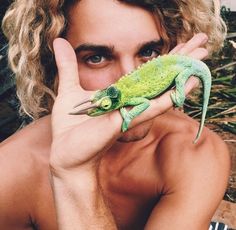 a man holding up a green lizard to his face while sitting on the ground in front of him