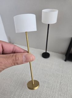 a hand holding a small white lamp next to a gold stand with a white shade on it