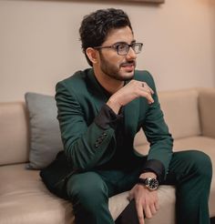 a man sitting on top of a couch wearing a green suit and black shirt with his hand under his chin