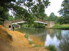 two people are walking on the side of a river near a bridge and some trees