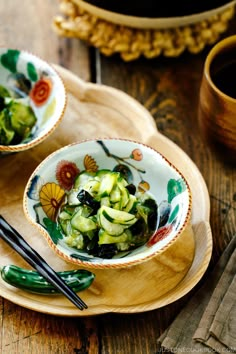 two bowls filled with cucumbers on top of a wooden tray next to chopsticks