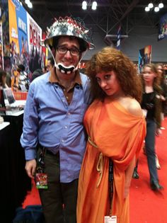 a man and woman in costumes posing for a photo at a convention or show with other people around them