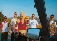 three men and a woman standing next to each other in front of a car mirror