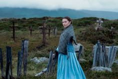 a woman in a blue dress standing next to a wooden fence on a hill with mountains in the background