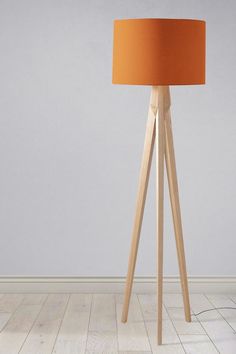 a floor lamp with an orange shade on the top and wooden legs, in front of a gray wall