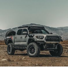 a gray truck parked on top of a dirt field next to some mountains in the background