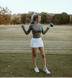 a woman holding a golf club on top of a grass covered field with trees in the background