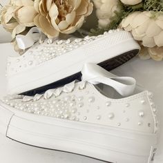 a pair of white shoes with pearls on them and flowers in the back ground next to it