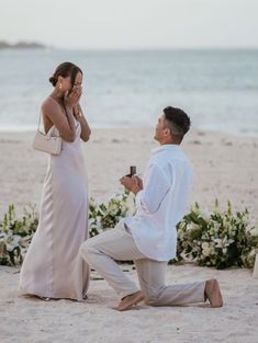 a man kneeling down next to a woman on the beach while holding a cell phone