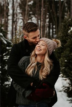 a man and woman standing in the snow hugging each other with trees in the background