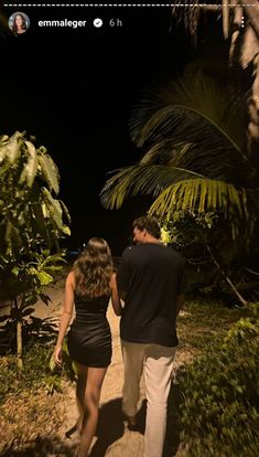 a man and woman walking down a dirt path at night with palm trees in the background