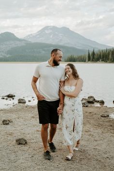 a man and woman standing next to each other near the water with mountains in the background