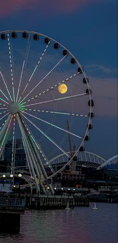 a large ferris wheel sitting next to the ocean under a full moon at night time