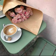 a cup of coffee and some pink flowers on a green table with a brown paper bag