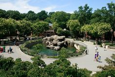 people are walking around in the park with trees and rocks on either side of the pond