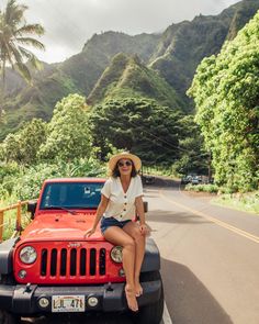 a woman sitting on the hood of a red jeep parked in front of a mountain