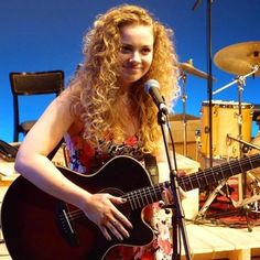 a woman holding a guitar and singing into a microphone while standing in front of a set of drums