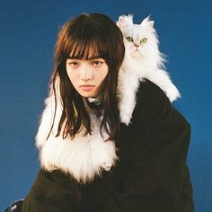 a woman holding a white cat on her shoulders and wearing a black coat over her shoulder