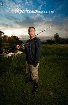 a man standing in the grass with a fishing pole and some water behind him at sunset