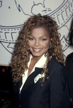 a woman with long curly hair standing in front of a sign and smiling at the camera