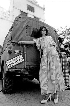 a black and white photo of a woman standing in front of a truck with her arm on the back of it