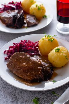 two white plates topped with meat, potatoes and red cabbage next to a glass of wine