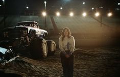 a woman standing next to an old truck in a dirt field with lights on the bleachers