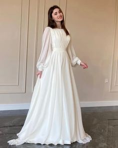 a woman wearing a white dress standing in front of a wall and posing for the camera