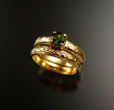 two gold wedding bands with a green stone in the center on a black surface,