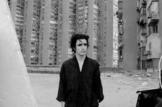 a black and white photo of a man standing in front of an unfinished building with his guitar