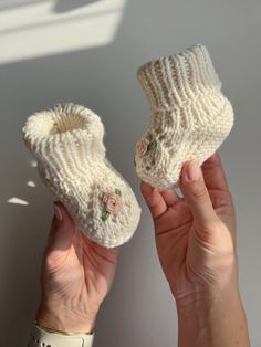 two hands holding up white knitted baby booties with flower appliques on them