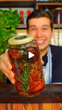 a man holding a jar filled with pickled tomatoes and herbs on top of a wooden table