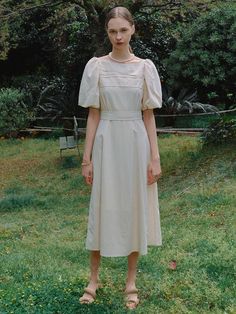 a woman standing in the grass wearing a white dress with short sleeves and an open neck